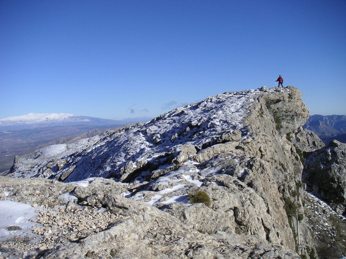 A lone hiker stands on a snow dusted limestone ridge in the Sierra Tejeda with a snowy distant range under a clear blue sky.