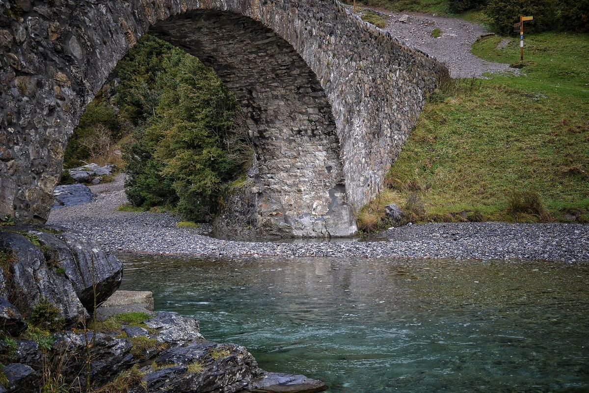 The old stone arch of the medieval bridge at Bujaruelo spans a clear turquoise river with grass and a trail signpost beside it.
