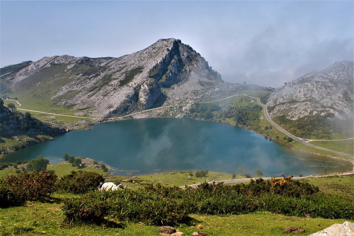 Lago Enol lies in a green bowl of limestone mountains in the Picos de Europa with a road curving along its shore and low cloud over the peaks.