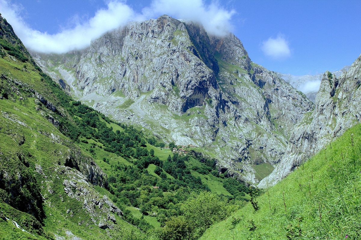 A steep green valley in the Picos de Europa with a tiny hamlet below a huge grey limestone wall under a blue sky with low cloud.