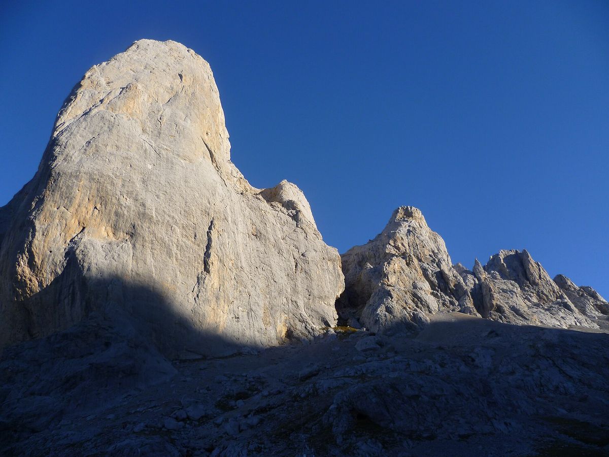 The pale limestone tower of Picu Urriellu, also called Naranjo de Bulnes, rises against a deep blue sky above rocky ground near the refuge.